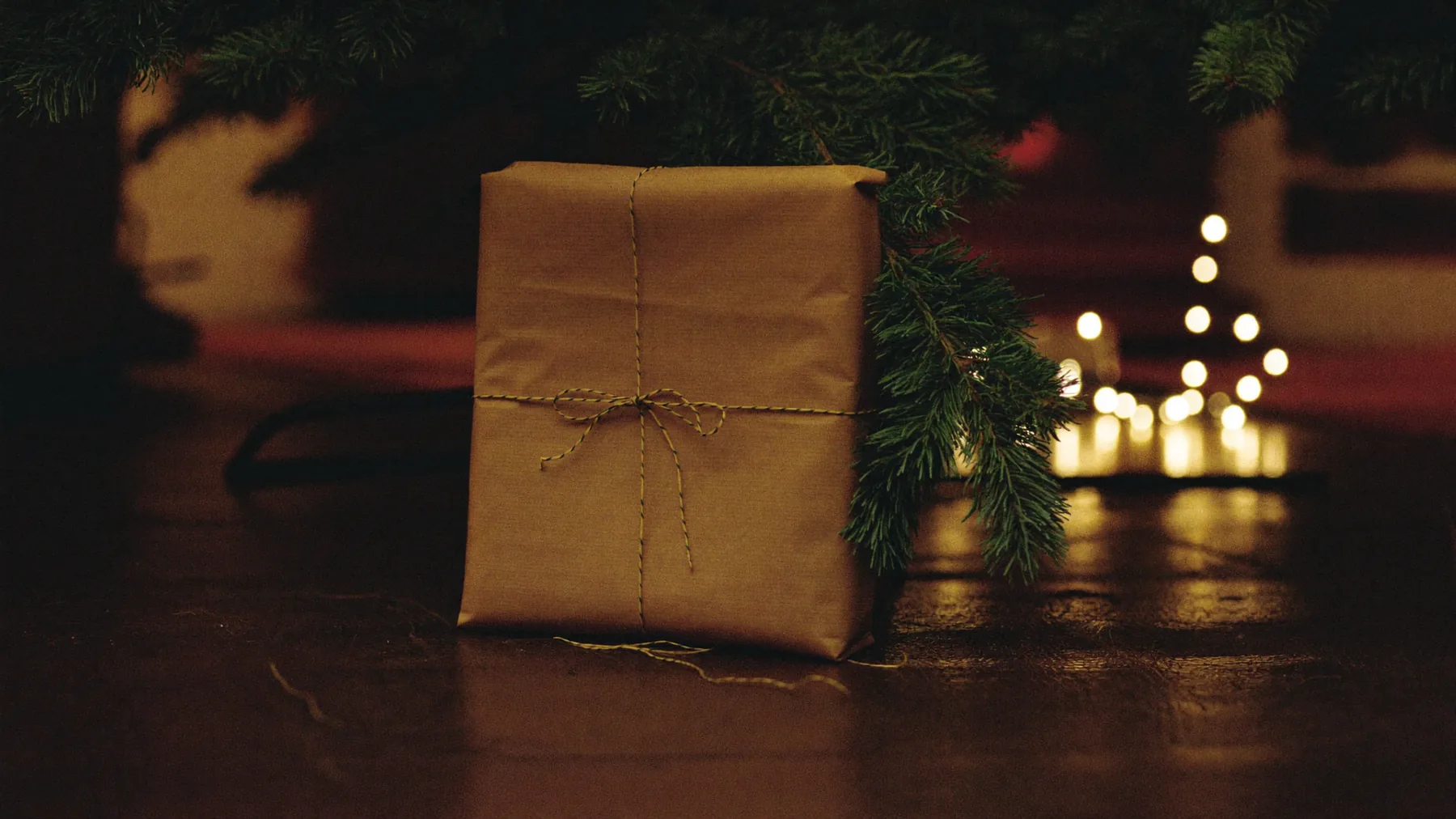 A flat parcel wrapped in brown paper and tied with twine, propped against a Christmas tree on a dark wooden floor with fairy lights out of focus behind it
